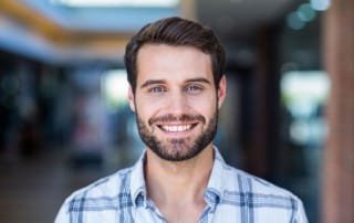 A younger man with short hair and a beard wearing a blue and white plaid button-up shirt and smiling