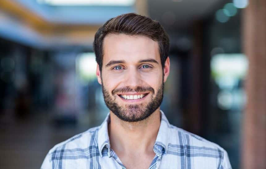 A younger man with short hair and a beard wearing a blue and white plaid button-up shirt and smiling